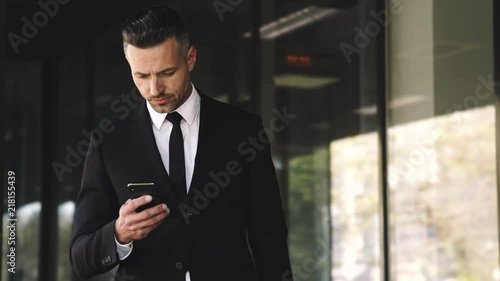 Serious business man dressed in suit using smartphone while waitng someone outside a glass building photo
