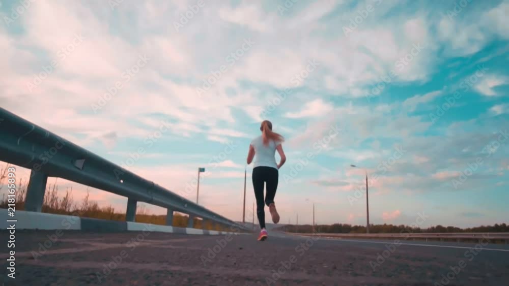 A sporty girl in a white t-shirt and with long hair runs along an empty track outside the city. The girl runs away into the distance, removed. Athletics classes in the setting sun. A sporty girl in a white t-shirt and with long hair runs along an empty track outside the city. The girl runs away into the distance, removed. Athletics classes in the setting sun.