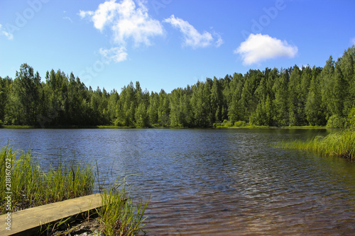 Summer, nature, ecology and landscape concept. Rest at the lake with clear water surrounded by forest. Blue sky with white clouds, wooden pier for fishing. Relax.