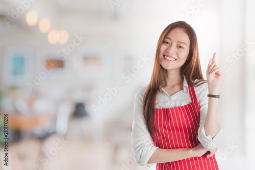 Portrait of Smiling beautiful young Asian woman wear red apron and hand pointing with copy space for your text or advertising on blurred cafe background.