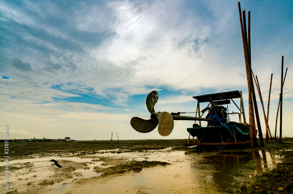 Fototapeta premium Blade boat on sea coast with blue sky and white clouds at boatyard. Propeller of longtail boat at dock hull. Dry bamboo pole embroidered on the mud beach.