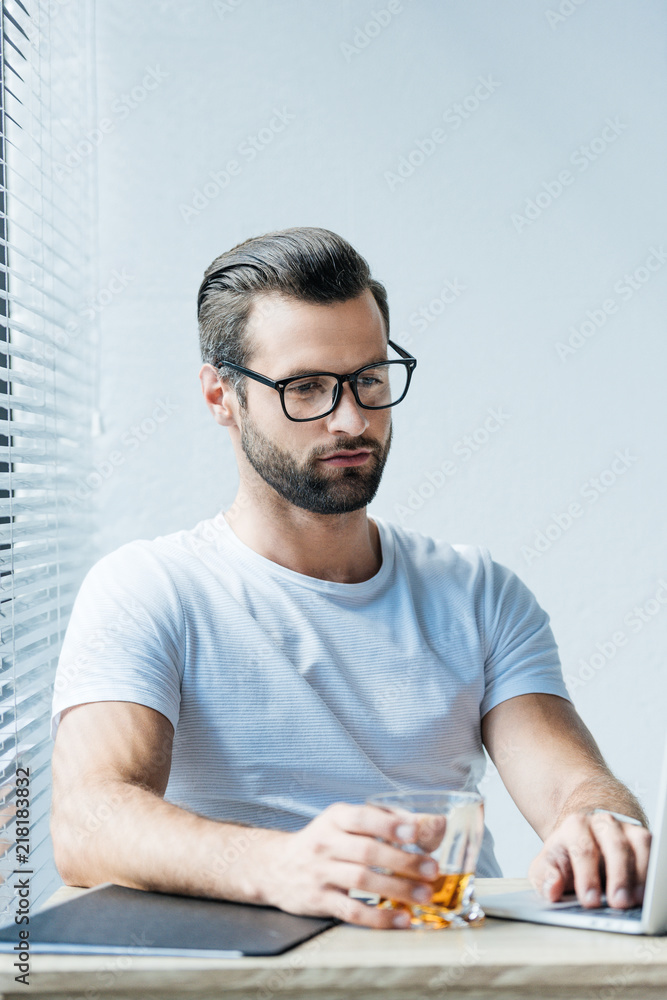bearded man in eyeglasses holding glass of whiskey and working with laptop bearded man in eyeglasses holding glass of whiskey and working with laptop