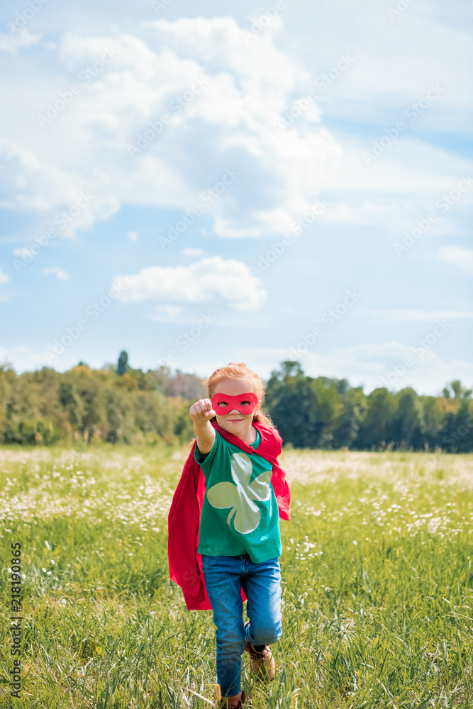 little kid in red superhero cape and mask with outstretched arm running in meadow on summer day little kid in red superhero cape and mask with outstretched arm running in meadow on summer day
