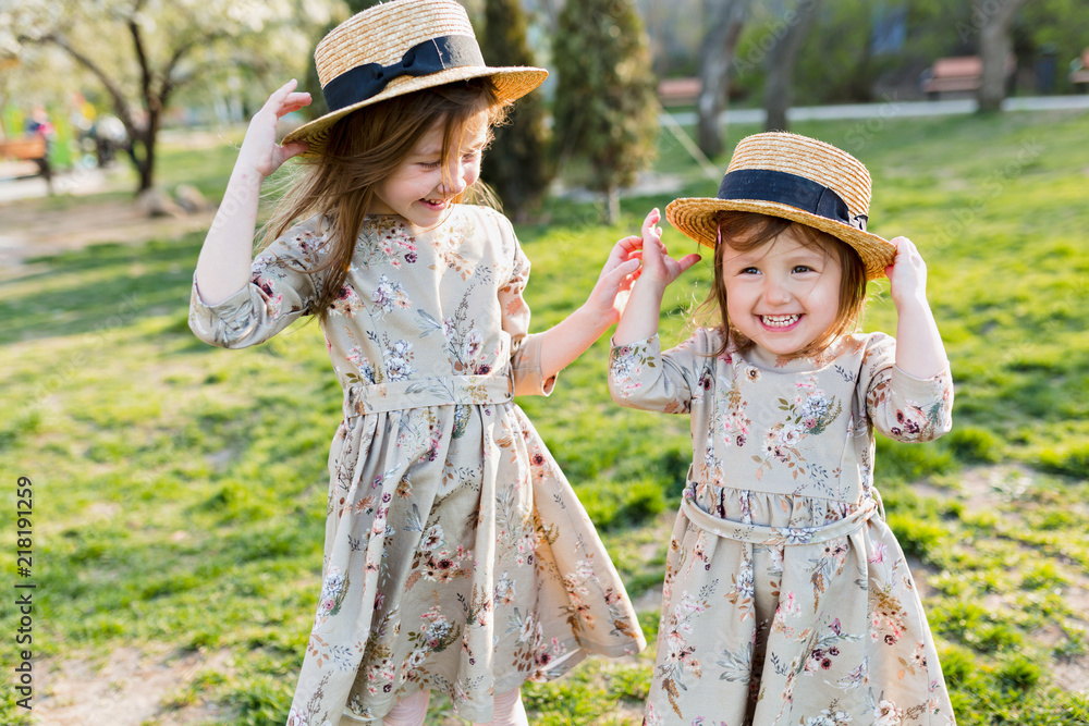 Incredible lovely happy two little girls dressed in similar summer dresses and hats plays outside in sunshine with happy true emotions. Two little sisters spending holidays in sunny summer park Stock ...
