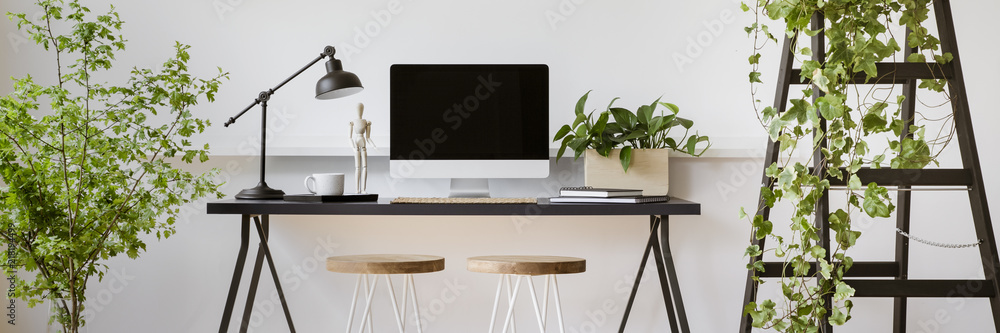 Real photo of two stools placed by home office desk with empty screen computer, lamp and coffee cup in bright room interior with fresh plants - obrazy, fototapety, plakaty 