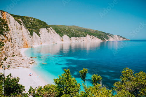 Sunny Fteri beach lagoon with rocky coastline, Kefalonia, Greece. Tourists under umbrella chill relax near clear blue emerald turquoise sea water photo