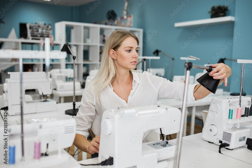 Obraz premium A smart-looking pretty blond woman wearing white shirt is sewing with the electric sewing-machine. Fashion, tailor's workshop.