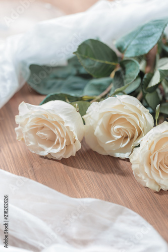Three white  beige roses lie on a wooden table. On the table lie roses and white transparent fabric. Wedding preparations.