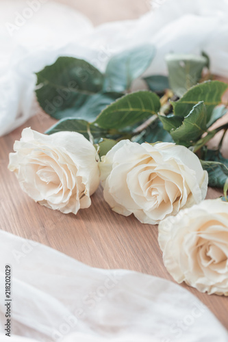 Three white, beige roses lie on a wooden table. On the table lie roses and white transparent fabric. Wedding preparations.