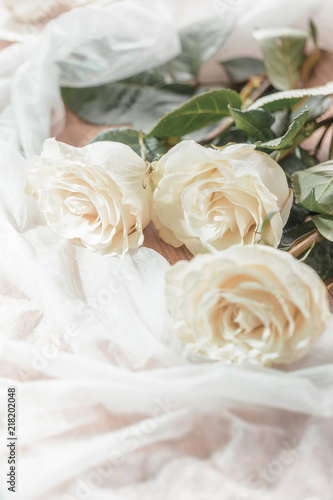 Three white  beige roses lie on a wooden table. On the table lie roses and white transparent fabric. Wedding preparations.