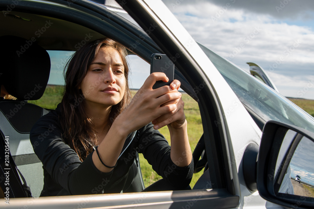 junge Frau sitzt in geparktem Auto und fotografiert mit Handy durch geöffnete Tür die Landschaft