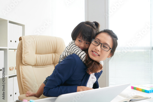 Adult Asian businesswoman in glasses sitting at table in office with adorable little girl on chair having fun