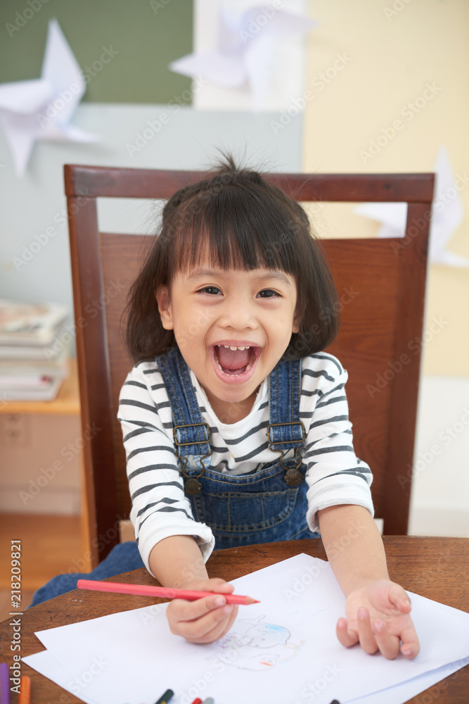 Charming Asian girl at table with paper and colorful pencils looking excitedly at camera Charming Asian girl at table with paper and colorful pencils looking excitedly at camera