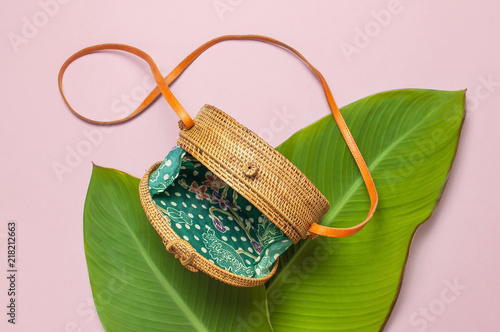 Fashionable handmade natural round rattan bag and tropical leaves on pink background flat lay. Top view with copy space. Trendy bamboo bag Ecobags from Bali. Summer fashion concept. photo