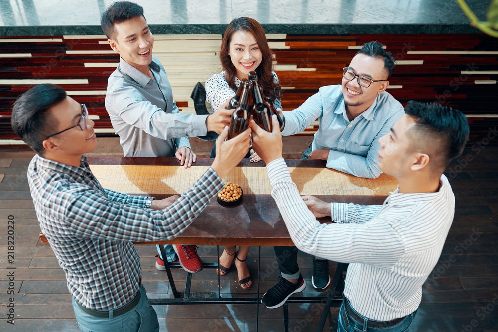 Obraz premium From above shot of Asian adult men and woman standing around wooden table in bar and clinking with beer bottles