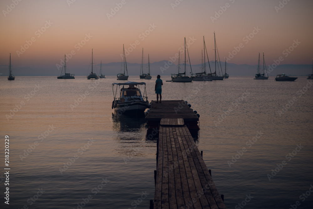 Man silhouette walking on the wooden pier on the sea bay with sailing boats and mountains on the background. Motot boat. Sunset over the horizon. Dusk Man silhouette walking on the wooden pier on the sea bay with sailing boats and mountains on the background. Motot boat. Sunset over the horizon. Dusk
