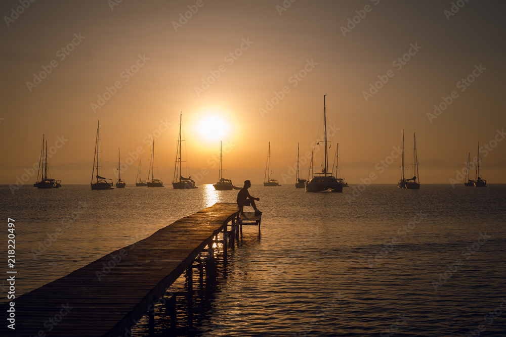 Obraz premium Man silhouette sitting on the wooden pier on the sea bay with sailing boats on the horizon. Breathtaking golden sunset.