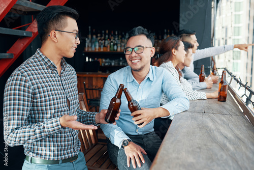 Asian men at bar counter toasting with beer bottles sitting with people on background in modern pub