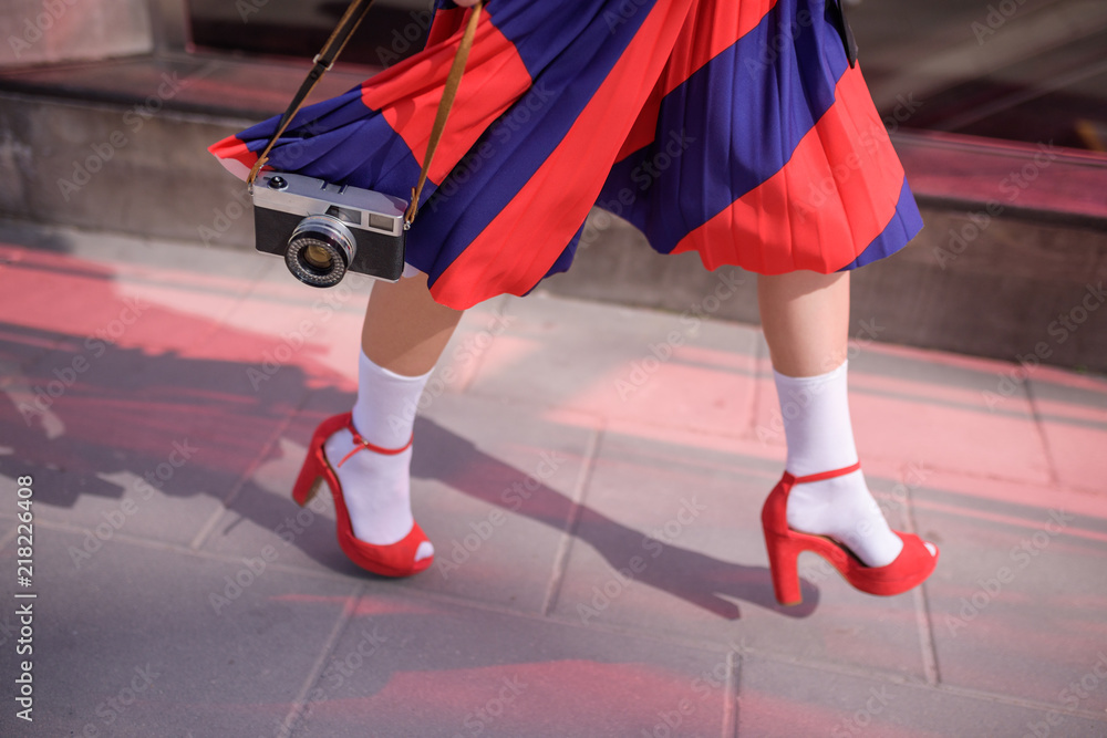 Elegant lady wearing stylish red skirt and shoes holds in hands vintage photo camera. Low angle view of her shapely legs. Selective focus Elegant lady wearing stylish red skirt and shoes holds in hands vintage photo camera. Low angle view of her shapely legs. Selective focus
