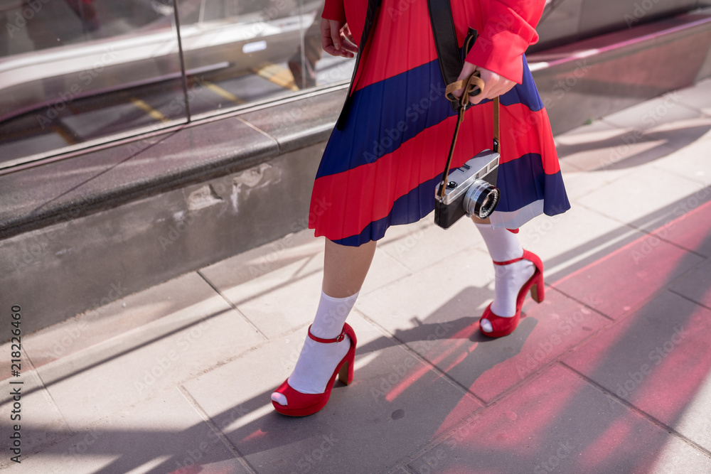 Elegant lady wearing stylish red skirt and shoes holds in hands vintage photo camera. Low angle view of her shapely legs. Selective focus Elegant lady wearing stylish red skirt and shoes holds in hands vintage photo camera. Low angle view of her shapely legs. Selective focus