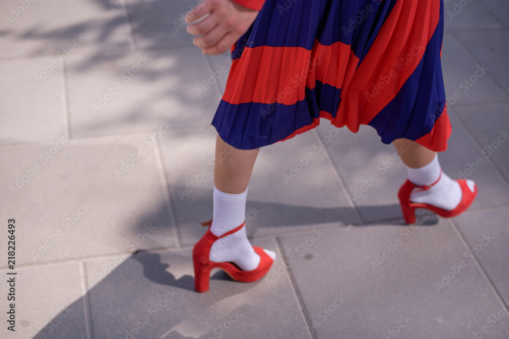 Young woman wearing stylish red skirt and shoes on an urban sidewalk. Low angle view of her shapely legs. Selective focus Young woman wearing stylish red skirt and shoes on an urban sidewalk. Low angle view of her shapely legs. Selective focus