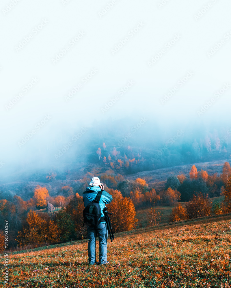 Naklejka premium Photographer taking photo of autumn landscape with foggy peaks and orange trees. Ukrainian Carpathians mountains