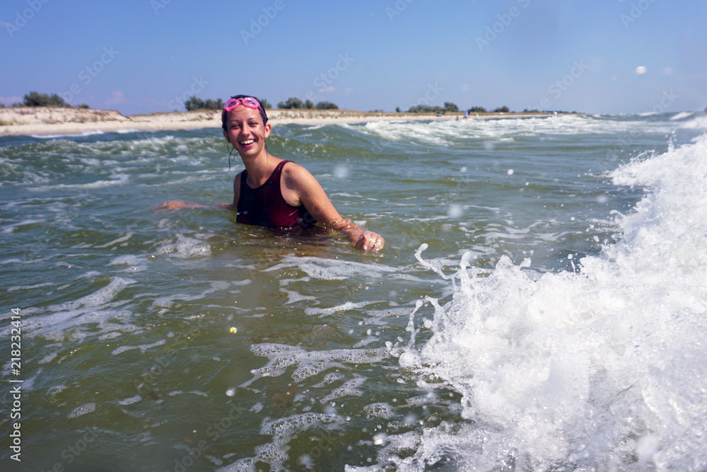 Young brunette girl in red swimsuit smiling with glasses for swimming in the sea, with beach island background and blue sky. Big wave with foam. Copyspace.