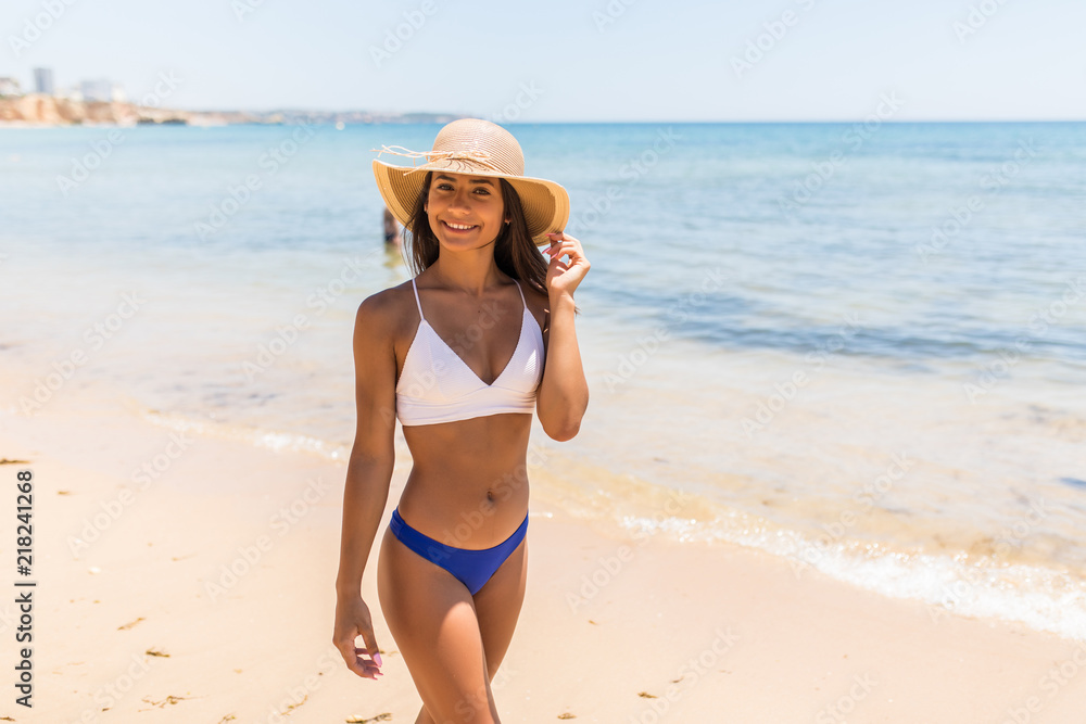 Blue sea, white sand paradise. Full length portrait of modern latin woman in bikini and beach straw hat on the seashore walking Blue sea, white sand paradise. Full length portrait of modern latin woman in bikini and beach straw hat on the seashore walking