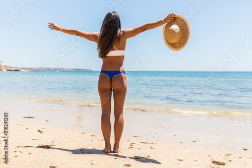 Woman on Beach standing with arms outstretched against turquoise sea. Rear view of female wearing bikini with raised hands. Carefree tourist is enjoying vacation at beach.