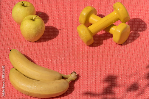Healthy lifestyle in modern colors: Two yellow dumbbells, apples, banans on the pink exercise matt. Shadows cast by sunlight on the surface.