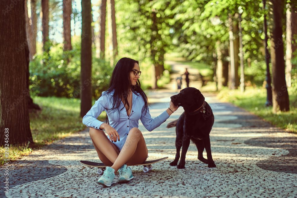 Happy brunette girl in glasses wearing a shirt and shorts sitting on a skateboard, walks with her dog at the park. Happy brunette girl in glasses wearing a shirt and shorts sitting on a skateboard, walks with her dog at the park.