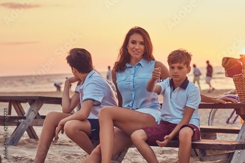 Happy family - mother with her sons sitting on a bench against the background of a seacoast at the bright sunset. © Fxquadro