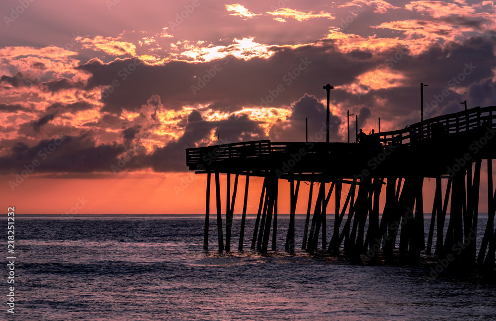 Dramatic red sky at sunrise that silhouettes a fishing pier and lights up the clouds. The ocean surface sparkles with the reflection of the morning light.