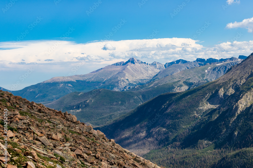 Sunny Colorado Rockies, with rocky hill in  left-front, dramatic  blue-grey mountains behind, with evergreens below. Bright blue sky  and clouds above.  Good for background, text.