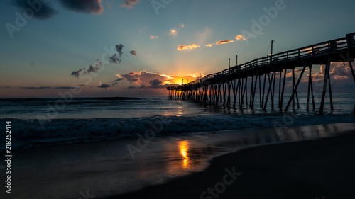 The morning sun rising behind the end of a wooden fishing pier extending out into the Atlantic Ocean. Sunlight reflected on the surface of the sea and the wet sand.