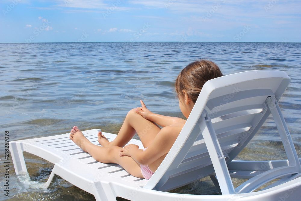 Little child on a deck chair on a sunny day, relaxing on the beach. Girl looking at the sea