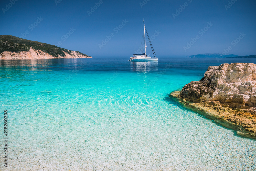 Beautiful azure blue lagoon with sailing catamaran yacht boat at anchor. Pure white pebble beach, some rocks in the sea