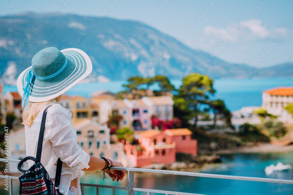 Tourist woman wearing blue sunhat and white clothes enjoying view of colorful tranquil village Assos on sunny day. Stylish female visiting Kefalonia in summer time on Greece travel vacation
