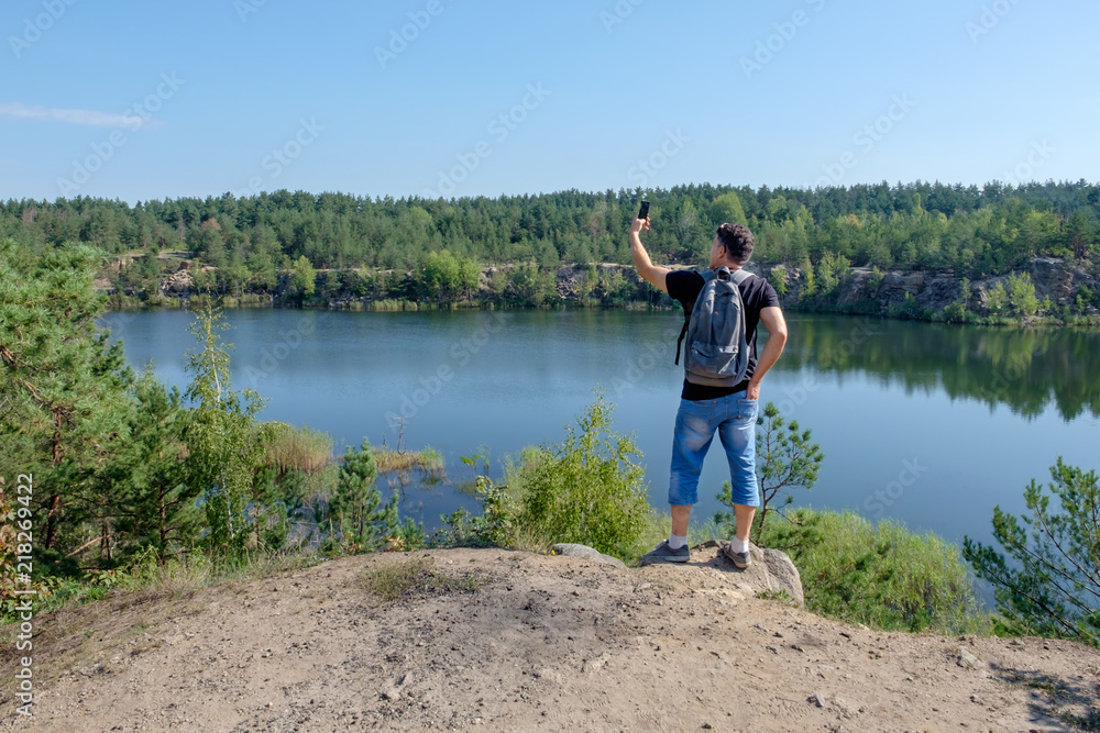 Senior white man with backpack stands on the rock and making selfie at the lake background.