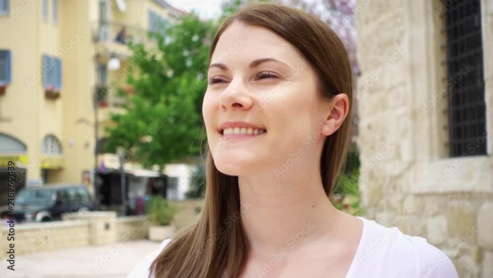 Smiling young woman in white t-shirt walking down street in european city in slow motion. Happy female looking around outside. Stone wall of old building and big green tree on background Smiling young woman in white t-shirt walking down street in european city in slow motion. Happy female looking around outside. Stone wall of old building and big green tree on background