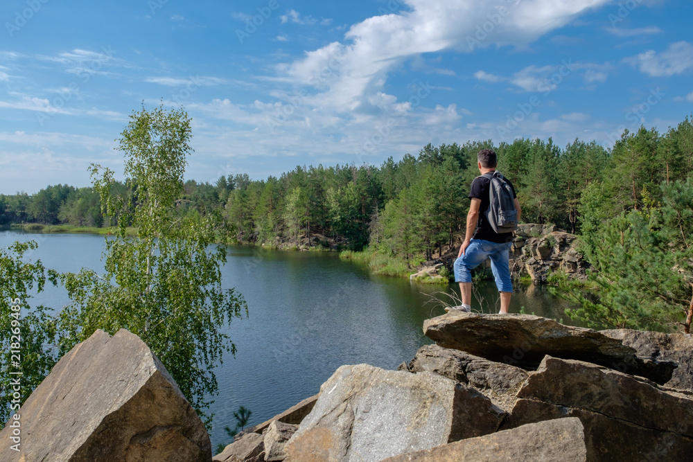 Adult white man with backpack stands on the rock and looks at the lake.