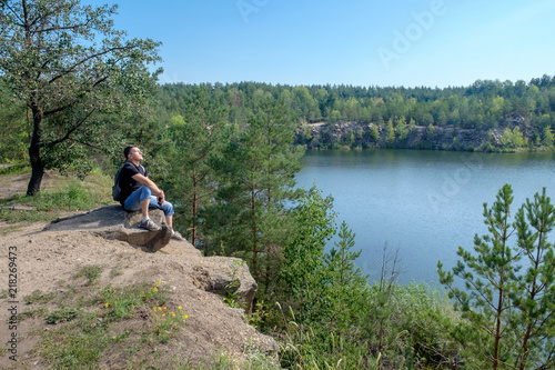 An senior white man with backpack sitting on the rock near the lake and keep a face to the sun. © Serhii