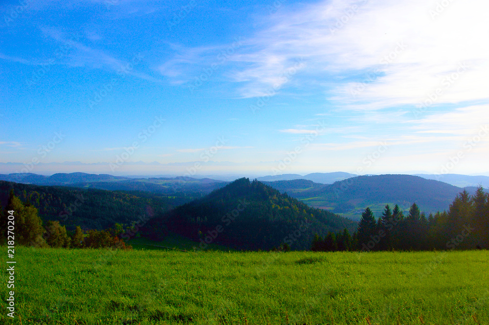 Hügelige Landschaft mit einem blauen Himmel und im Vordergrund eine grüne Wiese