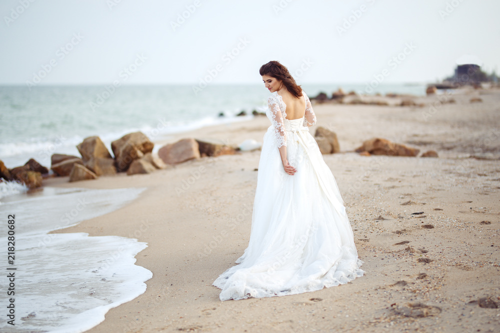 A beautiful bride in a white wedding dress is walking along the beach. Waves near her dress. Beautiful sunset atmosphere, summer evening. Wedding concept. A beautiful bride in a white wedding dress is walking along the beach. Waves near her dress. Beautiful sunset atmosphere, summer evening. Wedding concept.