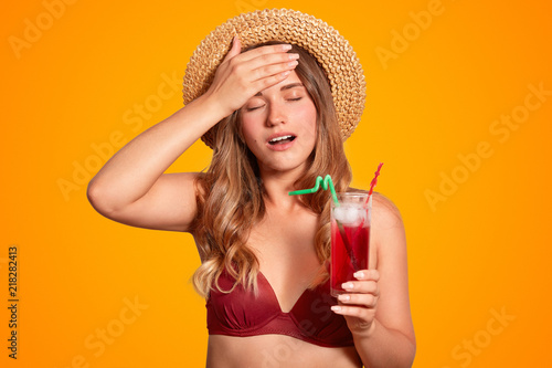 Tired female in bikini and summer hat, keeps hand on forehead, has headache after being outdoor during hot stuffy weather conditions, feels thirsty, drinks cold fresh cocktail to feel better