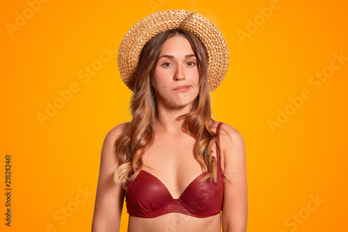 Photo of attractive young female cant stand stuff hot weather conditions during summer, blows air, feels sultry, dressed in swimwear and summer hat, isolated over orange background. Midday sun.
