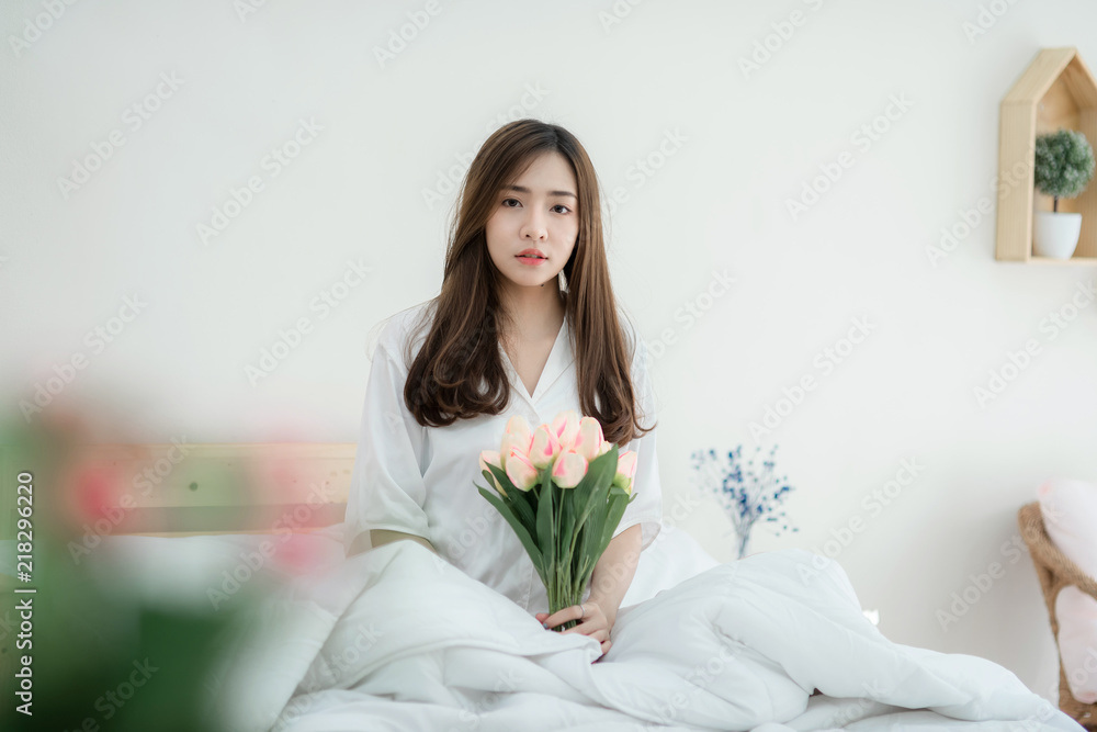 Beautiful young woman holding a bouquet of tulips siting on white bed. Happy morning. Portrait of a smiling pretty young lady relaxing in white bed. Beautiful young woman holding a bouquet of tulips siting on white bed. Happy morning. Portrait of a smiling pretty young lady relaxing in white bed.