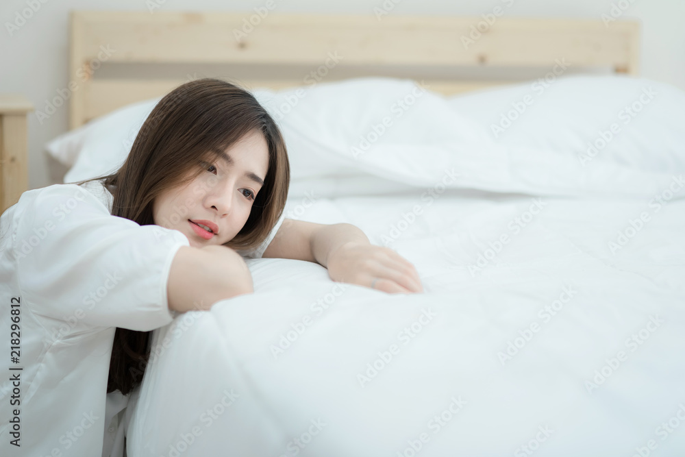Woman sitting lonely beside the bed in the white bedroom. Lazy Asian woman sitting beside the bed. Woman sitting lonely beside the bed in the white bedroom. Lazy Asian woman sitting beside the bed.