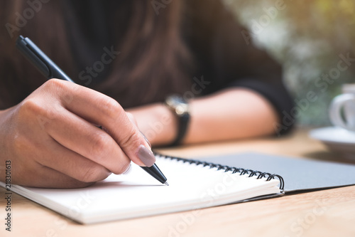 Closeup image of a woman's hand writing down on a white blank notebook with coffee cup on wooden table