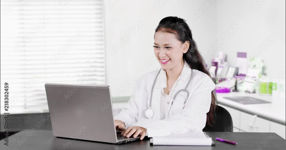 Young female doctor working with laptop computer on the table in the clinic and smiling at the camera. Shot in 4k resolution Young female doctor working with laptop computer on the table in the clinic and smiling at the camera. Shot in 4k resolution